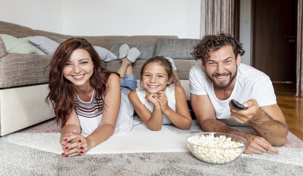 uśmiechnięta rodzina 2+1 leży na brzuchach na dywanie, ojciec trzyma pilot od telewizora w dłoni, na dywanie stoją pudełka z popcornem