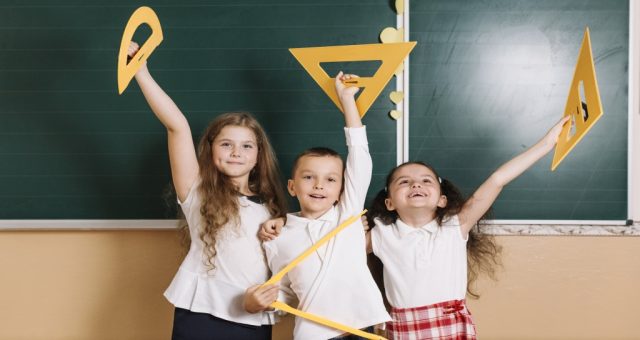 cieszące się dzieci na tle tablicy szkolnej, trzymające w rękach drewniane akcesoria do geometrii