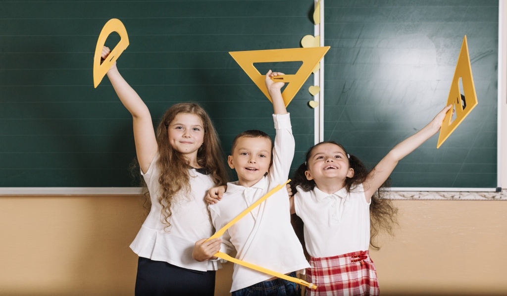 cieszące się dzieci na tle tablicy szkolnej, trzymające w rękach drewniane akcesoria do geometrii
