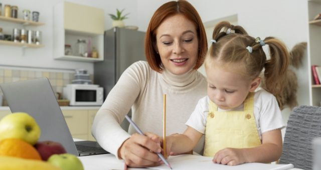Mama z córką przy stole, piszące coś w zeszycie ołówkiem