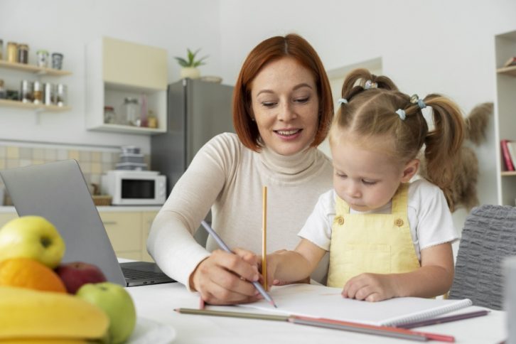 Mama z córką przy stole, piszące coś w zeszycie ołówkiem